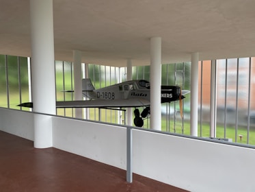A vintage airplane is suspended within a modern architectural space, featuring large windows and white pillars. The plane has a grey body with black and white markings, and a wooden propeller at the front. The background shows a lush green landscape through glass.