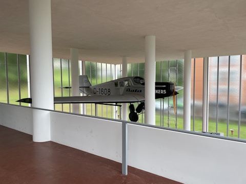 A vintage airplane is suspended within a modern architectural space, featuring large windows and white pillars. The plane has a grey body with black and white markings, and a wooden propeller at the front. The background shows a lush green landscape through glass.
