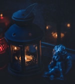A small candle lantern with star-shaped cutouts is lit, casting a warm glow in an otherwise dark environment. Beside the lantern is a small blue cherub statue. There are some distant lights or candles visible in the background, contributing to the overall dim and cozy atmosphere.