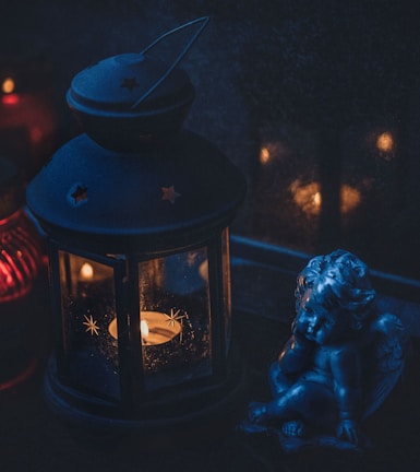 A small candle lantern with star-shaped cutouts is lit, casting a warm glow in an otherwise dark environment. Beside the lantern is a small blue cherub statue. There are some distant lights or candles visible in the background, contributing to the overall dim and cozy atmosphere.