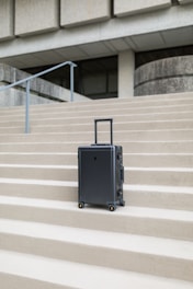 A sleek, modern suitcase with wheels stands on concrete steps in front of a large, austere, architectural structure. The suitcase is upright and positioned centrally, emphasizing its design against the geometric lines of the background.