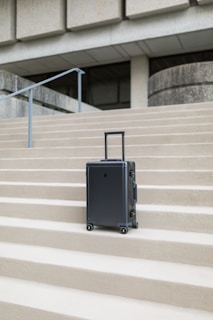 A sleek, modern suitcase with wheels stands on concrete steps in front of a large, austere, architectural structure. The suitcase is upright and positioned centrally, emphasizing its design against the geometric lines of the background.