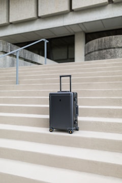 A sleek, modern suitcase with wheels stands on concrete steps in front of a large, austere, architectural structure. The suitcase is upright and positioned centrally, emphasizing its design against the geometric lines of the background.