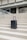 A sleek, modern suitcase with wheels stands on concrete steps in front of a large, austere, architectural structure. The suitcase is upright and positioned centrally, emphasizing its design against the geometric lines of the background.