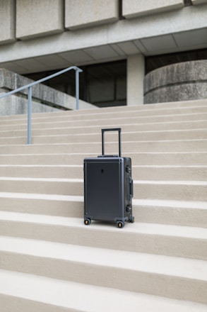 A sleek, modern suitcase with wheels stands on concrete steps in front of a large, austere, architectural structure. The suitcase is upright and positioned centrally, emphasizing its design against the geometric lines of the background.