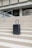 A sleek, modern suitcase with wheels stands on concrete steps in front of a large, austere, architectural structure. The suitcase is upright and positioned centrally, emphasizing its design against the geometric lines of the background.