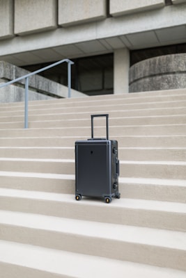 A sleek, modern suitcase with wheels stands on concrete steps in front of a large, austere, architectural structure. The suitcase is upright and positioned centrally, emphasizing its design against the geometric lines of the background.