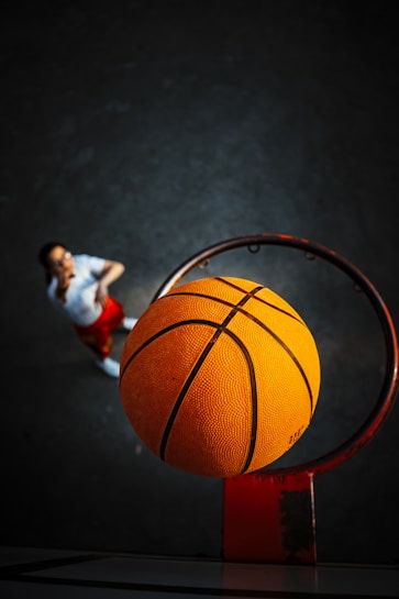 A basketball is captured mid-air directly above the hoop, about to enter. The focus is on the ball with the textured surface visible, while a player stands in the background on a dark court, looking up.