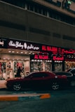 A street view featuring a shop front with illuminated signage in Arabic and English. The shop appears to sell watches, with a sign reading 'Watch Repairing.' A dark red car is parked in front of the store, and a person is walking past the entrance. The setting is urban with ambient street lighting.