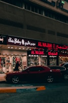 A street view featuring a shop front with illuminated signage in Arabic and English. The shop appears to sell watches, with a sign reading 'Watch Repairing.' A dark red car is parked in front of the store, and a person is walking past the entrance. The setting is urban with ambient street lighting.