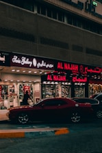 A street view featuring a shop front with illuminated signage in Arabic and English. The shop appears to sell watches, with a sign reading 'Watch Repairing.' A dark red car is parked in front of the store, and a person is walking past the entrance. The setting is urban with ambient street lighting.