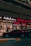 A street view featuring a shop front with illuminated signage in Arabic and English. The shop appears to sell watches, with a sign reading 'Watch Repairing.' A dark red car is parked in front of the store, and a person is walking past the entrance. The setting is urban with ambient street lighting.