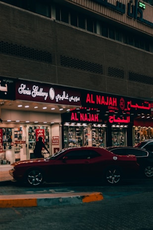 A street view featuring a shop front with illuminated signage in Arabic and English. The shop appears to sell watches, with a sign reading 'Watch Repairing.' A dark red car is parked in front of the store, and a person is walking past the entrance. The setting is urban with ambient street lighting.