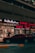 A street view featuring a shop front with illuminated signage in Arabic and English. The shop appears to sell watches, with a sign reading 'Watch Repairing.' A dark red car is parked in front of the store, and a person is walking past the entrance. The setting is urban with ambient street lighting.