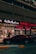 A street view featuring a shop front with illuminated signage in Arabic and English. The shop appears to sell watches, with a sign reading 'Watch Repairing.' A dark red car is parked in front of the store, and a person is walking past the entrance. The setting is urban with ambient street lighting.