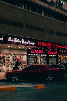 A street view featuring a shop front with illuminated signage in Arabic and English. The shop appears to sell watches, with a sign reading 'Watch Repairing.' A dark red car is parked in front of the store, and a person is walking past the entrance. The setting is urban with ambient street lighting.