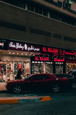 A street view featuring a shop front with illuminated signage in Arabic and English. The shop appears to sell watches, with a sign reading 'Watch Repairing.' A dark red car is parked in front of the store, and a person is walking past the entrance. The setting is urban with ambient street lighting.