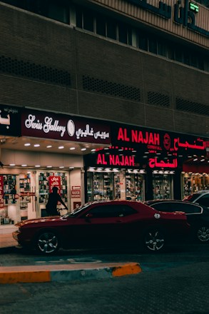 A street view featuring a shop front with illuminated signage in Arabic and English. The shop appears to sell watches, with a sign reading 'Watch Repairing.' A dark red car is parked in front of the store, and a person is walking past the entrance. The setting is urban with ambient street lighting.