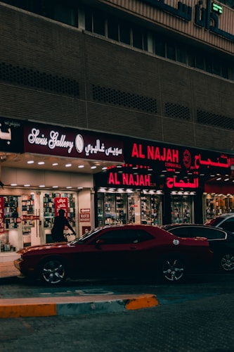 A street view featuring a shop front with illuminated signage in Arabic and English. The shop appears to sell watches, with a sign reading 'Watch Repairing.' A dark red car is parked in front of the store, and a person is walking past the entrance. The setting is urban with ambient street lighting.