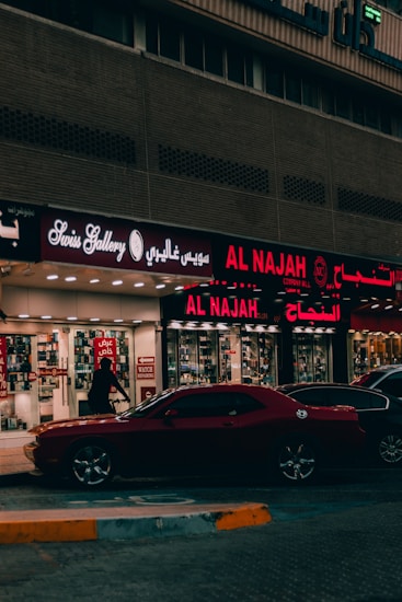 A street view featuring a shop front with illuminated signage in Arabic and English. The shop appears to sell watches, with a sign reading 'Watch Repairing.' A dark red car is parked in front of the store, and a person is walking past the entrance. The setting is urban with ambient street lighting.