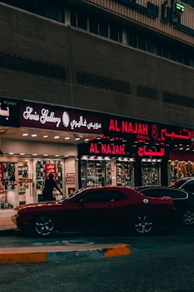 A street view featuring a shop front with illuminated signage in Arabic and English. The shop appears to sell watches, with a sign reading 'Watch Repairing.' A dark red car is parked in front of the store, and a person is walking past the entrance. The setting is urban with ambient street lighting.