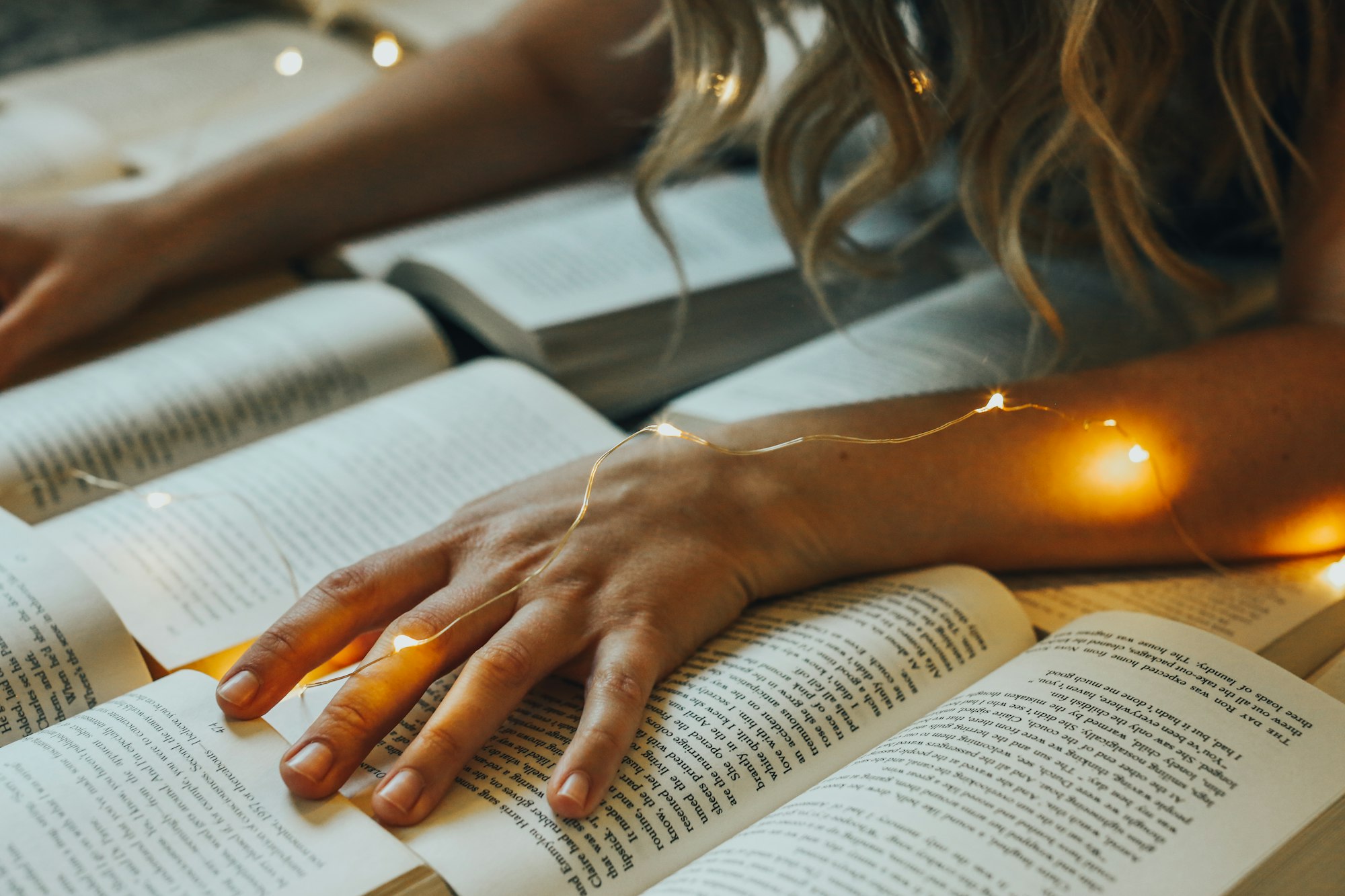 A girl engrossed in reading multiple books with delicate fairy lights illuminating her study area.