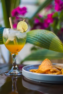 A refreshing drink garnished with lime and cinnamon sticks is placed on a polished surface next to a plate filled with folded tortilla chips. In the background, vibrant pink bougainvillea flowers and large green leaves add a tropical vibe.
