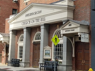 A brick building featuring classical architectural elements with columns and arched windows. The sign reads 'Swedenborgian Church on the Hill' above an entrance that has '140 Bowdoin Street.' Nearby is a yellow pedestrian crossing sign, two benches, and a small restaurant sign that says 'Fill-A-Buster.'