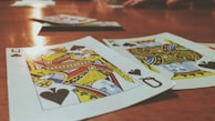 Playing cards are spread out on a wooden table, with the Queen and King of Spades prominently visible. The focus is on the vibrant colors and detailed designs of the cards, while other cards and hands are slightly blurred in the background, suggesting a game in progress.