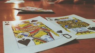Playing cards are spread out on a wooden table, with the Queen and King of Spades prominently visible. The focus is on the vibrant colors and detailed designs of the cards, while other cards and hands are slightly blurred in the background, suggesting a game in progress.