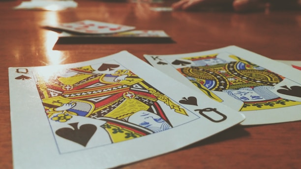 Playing cards are spread out on a wooden table, with the Queen and King of Spades prominently visible. The focus is on the vibrant colors and detailed designs of the cards, while other cards and hands are slightly blurred in the background, suggesting a game in progress.