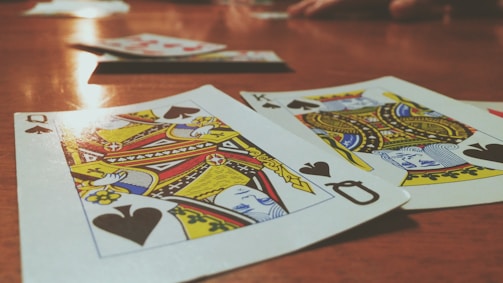 Playing cards are spread out on a wooden table, with the Queen and King of Spades prominently visible. The focus is on the vibrant colors and detailed designs of the cards, while other cards and hands are slightly blurred in the background, suggesting a game in progress.