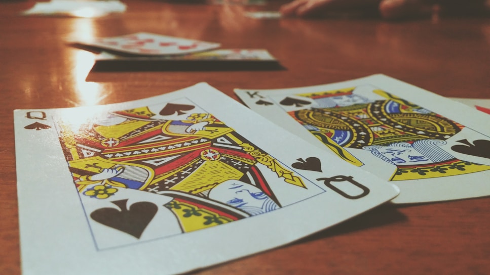 Playing cards are spread out on a wooden table, with the Queen and King of Spades prominently visible. The focus is on the vibrant colors and detailed designs of the cards, while other cards and hands are slightly blurred in the background, suggesting a game in progress.