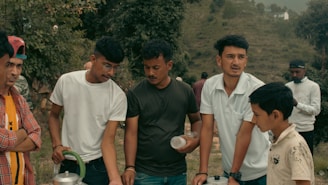 A group of young people is gathered outdoors in a rural area, surrounded by greenery. One person is holding a metallic container, and another holds a transparent plastic cup. They appear to be engaged in a conversation.