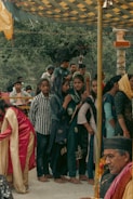 A group of people, mainly women dressed in traditional attire, are gathered under a decorated canopy. Some women wear headscarves and seem to be engaged in a cultural or religious event. A man, possibly an elder, is seated nearby with garlands around his neck, enhancing the traditional atmosphere.