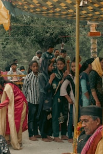 A group of people, mainly women dressed in traditional attire, are gathered under a decorated canopy. Some women wear headscarves and seem to be engaged in a cultural or religious event. A man, possibly an elder, is seated nearby with garlands around his neck, enhancing the traditional atmosphere.