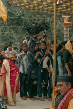 A group of people, mainly women dressed in traditional attire, are gathered under a decorated canopy. Some women wear headscarves and seem to be engaged in a cultural or religious event. A man, possibly an elder, is seated nearby with garlands around his neck, enhancing the traditional atmosphere.