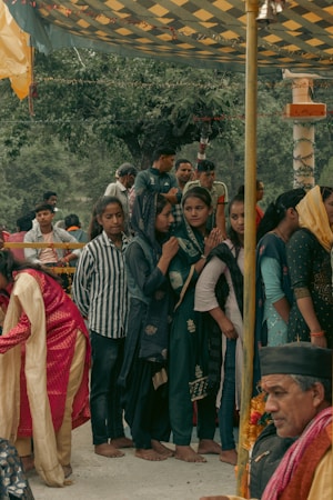 A group of people, mainly women dressed in traditional attire, are gathered under a decorated canopy. Some women wear headscarves and seem to be engaged in a cultural or religious event. A man, possibly an elder, is seated nearby with garlands around his neck, enhancing the traditional atmosphere.