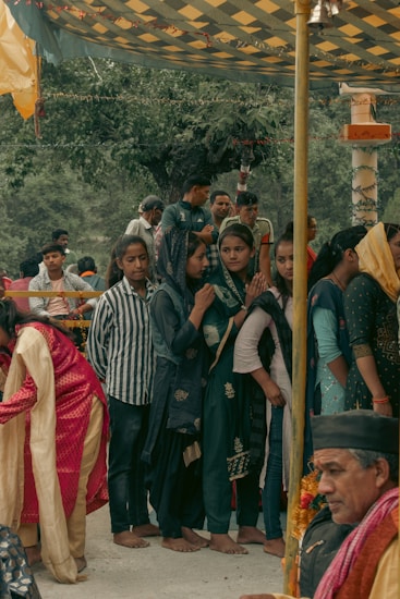 A group of people, mainly women dressed in traditional attire, are gathered under a decorated canopy. Some women wear headscarves and seem to be engaged in a cultural or religious event. A man, possibly an elder, is seated nearby with garlands around his neck, enhancing the traditional atmosphere.