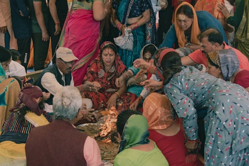 A group of people gathered closely together performing a traditional ritual around a small fire. Many individuals are dressed in colorful traditional clothing, displaying patterns and bright fabrics. The central figure is a woman in red attire, surrounded by others who are engaging in the ritual. Some individuals have their hair covered, and a few wear jewelry, indicating a cultural or ceremonial setting.