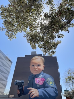 A large mural on a building depicts a baby dressed in a police officer's uniform, holding a toy gun. The baby has a serious expression and wears a pink toy camera. Trees and bright sunlight are visible above the mural, partially obscuring the sky.