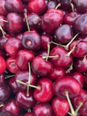 A close-up view of a pile of ripe red cherries with green stems attached. The cherries are glossy and appear fresh, with a rich red color that varies slightly among the individual fruits. The image captures the natural texture and juiciness of the cherries.