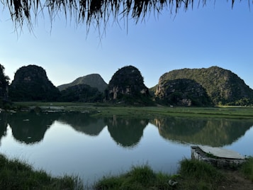 A serene landscape features towering, lush green limestone mountains reflected in a calm river or lake. The surface of the water is smooth, mirroring the shapes and textures of the cliffs. In the foreground, there is a grassy area with a stone platform, and a thatched roof can be seen at the top, suggesting a shaded viewpoint.