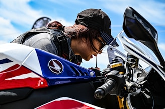 A person wearing a cap and sunglasses leans close to a motorcycle, appearing focused on a task. The motorcycle features a prominent Honda emblem and has a design with red, white, and blue colors. A clear blue sky is visible in the background.