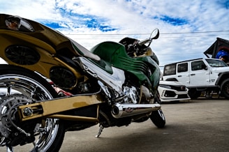 A uniquely designed motorcycle with a metallic gold frame and green accents appears in the foreground. The bike features prominent chrome details and a sleek, aerodynamic shape. In the background, a white Jeep and another sporty vehicle are parked, set against a partly cloudy blue sky.