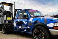 A blue pickup truck with a geometric camouflage pattern is parked next to a diesel fuel pump. The truck has custom stickers and decals on the windshield and body. The fuel pump is labeled 'Marked Diesel'.