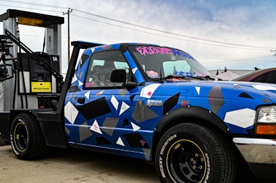 A blue pickup truck with a geometric camouflage pattern is parked next to a diesel fuel pump. The truck has custom stickers and decals on the windshield and body. The fuel pump is labeled 'Marked Diesel'.
