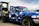 A blue pickup truck with a geometric camouflage pattern is parked next to a diesel fuel pump. The truck has custom stickers and decals on the windshield and body. The fuel pump is labeled 'Marked Diesel'.