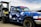 A blue pickup truck with a geometric camouflage pattern is parked next to a diesel fuel pump. The truck has custom stickers and decals on the windshield and body. The fuel pump is labeled 'Marked Diesel'.