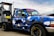 A blue pickup truck with a geometric camouflage pattern is parked next to a diesel fuel pump. The truck has custom stickers and decals on the windshield and body. The fuel pump is labeled 'Marked Diesel'.